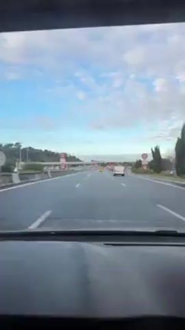 The video shows a view from inside a car driving on a highway. The sky is partly cloudy with a blue hue.  There are multiple lanes on the highway, and other cars are visible, including a white van. There are overhead signs and speed limit signs indicating 70. A toll booth structure is visible in the distance with red lights illuminated.  The dashboard of the car is visible in the foreground.