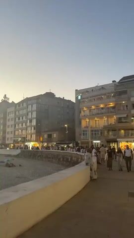The video begins with a view of a beach promenade at sunset. Buildings line the promenade, and the sky is a gradient of orange and blue. People are visible walking along the promenade and relaxing on the beach. The camera then pans to the left, showing a wider view of the beach with the ocean and the horizon. The sunset continues to color the sky with warm hues. The video captures the serene atmosphere of a coastal town during the evening.