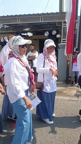 The video shows a group of women dressed in white shirts, blue skirts, and white headscarves in what appears to be a parade or street event. Some of the women are wearing white baseball caps with the word "SPORT" on them, and sunglasses. They are smiling, dancing, and gesturing. One woman in the foreground is giving a thumbs-up. In the background, there are houses and trees, suggesting an outdoor, residential setting. A flag that appears to be the Indonesian flag is visible in the upper left corner. There are decorative elements hanging from wires overhead. The lighting suggests it is daytime and sunny.