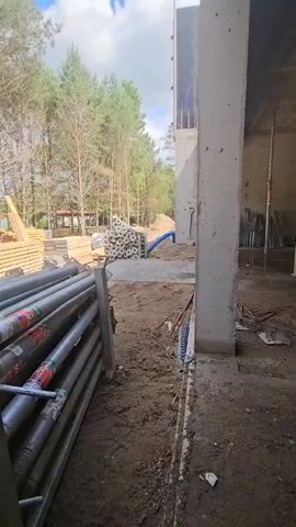 The video shows a construction site with stacks of metal poles in the foreground. In the background, there are piles of lumber and trees. The ground is dirt. The scene appears to be outdoors on a sunny day.