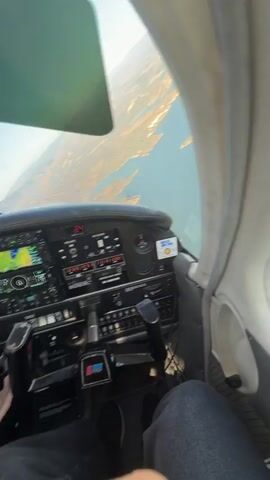 The video begins from the perspective of someone in the cockpit of a small airplane, looking out the window at a body of water and mountainous terrain. The camera then shifts to a view of the airplane's dashboard, which is filled with various instruments, screens, and controls. One of the screens displays navigation information. A person's hands are visible on the controls. The camera then pans back to the view out the window, showing the airplane banking to the right over the water and landscape. The video is shot during the daytime, with clear skies and sunlight.