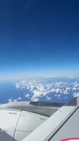 The view from an airplane window shows a bright blue sky with scattered white clouds and a body of water below. The plane's wing, with the words "NO STEP" printed on it, is visible in the foreground. The camera is still, and there are no visible people or specific actions.
