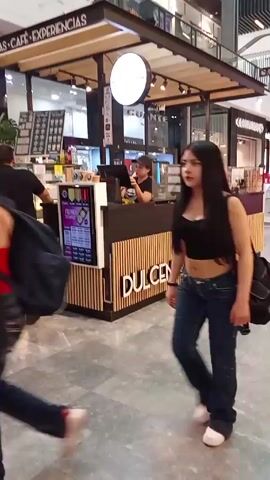 The video shows the inside of a shopping mall, focusing on a coffee and donut kiosk called "DUL CENTORNO".  People are seen walking past the kiosk. Some patrons are being served at the counter by an employee wearing a cap. The kiosk has displays for coffee, donuts and "EXPERIENCIAS".  The lighting is bright, and the floor is made of gray tiles.