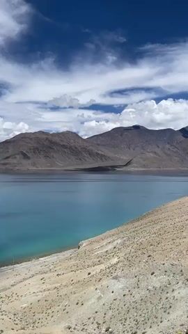 A wide shot captures a serene lake with turquoise and deep blue waters, bordered by a sandy shore on the right and barren, mountainous terrain on the left. The sky above is a vibrant blue, adorned with scattered white clouds. The camera pans slowly to the left, revealing more of the vast expanse of the lake and the surrounding rugged, brown mountains. The water's surface is calm, reflecting the sky and mountains. The landscape appears arid and sparsely vegetated.