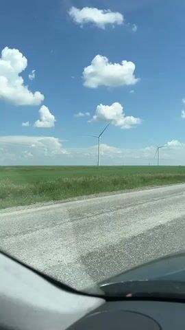 The media file shows a view from inside a vehicle traveling on a dusty, rural road. The road is made of gravel and is rough. On the right side of the road, there is a white van with a license plate that reads "KZ YUD 819". In the distance, a vast green field stretches to the horizon, dotted with numerous tall white wind turbines. The sky is a bright blue, filled with fluffy white clouds. The sun appears to be high in the sky, suggesting it is daytime, likely midday. The scene evokes a sense of open space and possibly a journey through a rural, agricultural landscape in Kazakhstan. The wind turbines indicate a location focused on renewable energy. There are no people visible within the frame, only the objects and the landscape.