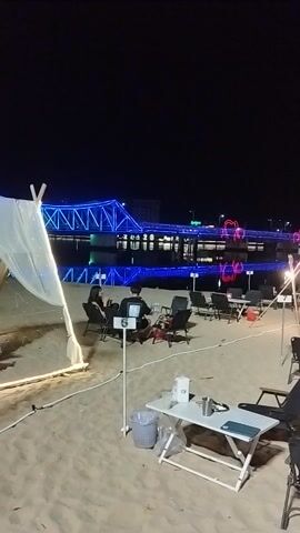 This video captures a lively night scene on a beach in Kampot, Cambodia. The camera pans across a sandy expanse dotted with numerous tables and chairs, suggesting an outdoor dining or entertainment area. String lights, some with heart-shaped accents, and bright blue neon lights illuminate the scene, creating a festive atmosphere. 

Several people are visible, either seated at the tables or moving about, though their faces are not clearly discernible. They appear to be enjoying the evening, with the overall mood seeming relaxed and convivial. In the background, illuminated buildings and what appears to be a Ferris wheel or similar amusement ride add to the visual spectacle. The dark sky indicates that it is nighttime, and the presence of beach furniture implies a location by the sea. 

A large sign with text in Khmer script is prominently displayed in the foreground, though the specific content is not legible. Other objects in the scene include tables, chairs, a red trash bin, and various poles and stands, likely for lighting or decorations. The sandy ground and the overall setup suggest a casual beachfront establishment. There is no clear indication of specific weather conditions beyond it being dark. The video seems to be documenting a night out at a popular beachside venue in Kampot.