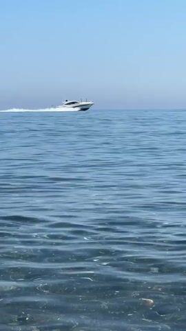 A sleek, white motor yacht speeds across a calm, blue ocean under a clear, bright sky, suggesting it's a sunny day. The yacht is positioned on the right side of the frame, leaving a white wake trailing behind it. The water in the foreground is clear and shallow, revealing a rocky or pebble-strewn seabed. In the distance, a small yellow buoy is visible, hinting at a marked area or perhaps a navigational marker. The scene evokes a sense of leisure and speed, characteristic of a warm, seaside environment. The location is identified as El Velerín, Spain, which aligns with the Mediterranean aesthetic. There is no visible text in the media file.