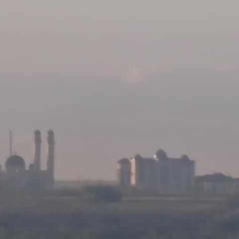 The video shows a wide shot of an Islamic mosque with minarets and a dome, as well as another building with domes, in the distance. The buildings appear to be situated in a flat landscape with some trees in the foreground. The sky is overcast with a hazy atmosphere. The video is grainy and out of focus, suggesting it was filmed from a distance or with a low-quality camera.