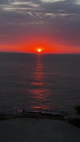A vertical video captures a vibrant sunset over a calm body of water. The sun, a bright orange orb, is positioned near the horizon, casting a shimmering, reddish-orange reflection that stretches across the water's surface. The sky above the horizon is a gradient of intense red and orange, transitioning to cooler tones of purple and gray with scattered clouds towards the top of the frame. In the foreground, a dark, paved area is visible, with a low railing and posts, suggesting a viewing platform or edge. To the right, a silhouette of a tree or foliage can be seen against the sky. The overall mood is serene and picturesque, highlighting the natural beauty of the sunset.