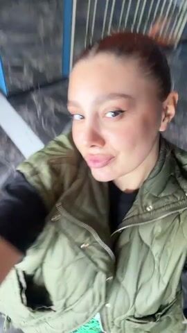 A woman in a green vest puckers her lips toward the camera in a selfie video. She stands indoors on what appears to be a marble floor. Her hair is pulled back, and she wears visible makeup. The camera rotates slightly as she maintains her pose.  Background details show a partial view of a white metal barrier. Loud music is audible in the background.
