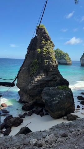 The video shows a large, dark rock formation on a white sand beach, partially surrounded by turquoise water. The rock is covered with some greenery. In the background, another smaller, green island sits in the ocean under a clear blue sky with a few scattered clouds. A black cable or rope hangs from the top left of the frame, suggesting a structure nearby. The sea gently laps against the shore.