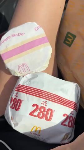 The video opens with a close-up shot of two McDonald's burgers wrapped in paper. The burger in the foreground is larger and wrapped in white paper with red stripes and the text "Le 280" printed repeatedly in red. The burger in the background is smaller and wrapped in white paper with pink and yellow stripes and the text "Croque McDo" and the McDonald's logo printed on it. A McDonald's paper bag is visible to the right of the burgers. A hand then enters the frame and holds up a receipt. The receipt shows "2 PRODUITS", "1 280 Original", and "1 Croque". It also indicates "DRIVE THRU 102" and a date and time stamp of "24/08/2023 11:21:30". The hand moves the receipt to reveal the two burgers more clearly. The scene appears to be outdoors, likely in a car, given the natural lighting and the presence of a person's leg.