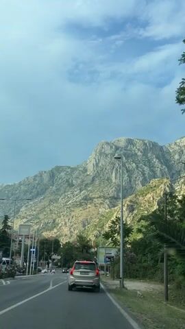 A street view with a mountain backdrop. A silver Volvo SUV is driving on the road, with other vehicles and pedestrians visible in the background. The sky is cloudy with patches of blue. The time stamp indicates the video begins at 00:00.