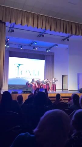 A dance group of around 10 young women is performing on stage. They are wearing grey shirts and maroon pants. The stage has a large screen behind them with the logo of a "Tema" acrobatic dance club. The lighting is blue and there are several spotlights on the stage. In the foreground, we see the heads of people in the audience, with one person holding up a phone to record the performance. The theater has brown curtains and blue and green stage lights. The room is dimly lit, focusing attention on the stage.