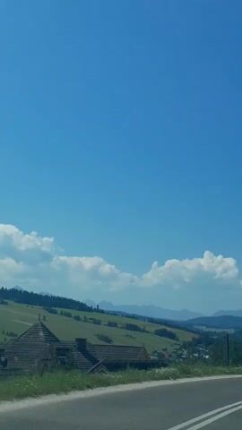 The video shows a wide blue sky with some white clouds. Below the sky, there is a green hillside with some trees and houses. In the foreground, there is a road with white lines.