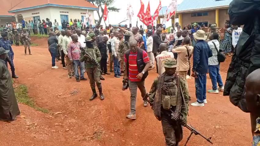 The video shows a large crowd gathered on a dirt road in front of two buildings. Many individuals in the crowd are wearing civilian clothing while others are in military attire, carrying weapons. Several people are holding red flags with white designs. Some people in the crowd are taking photos or videos with their phones or cameras. Individuals wearing "PRESS" vests are seen among the crowd.  The environment appears to be rural, and the buildings have a basic construction.