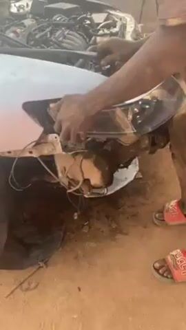 A person wearing a light brown t-shirt, dark pants, and red Gucci slides is installing a headlight on a white car. The car’s hood is open and the engine is visible. The front bumper is missing and the surrounding area is dirty. The person’s hands are soiled, suggesting they have been working on the car for some time. The car is outdoors on a dirt or gravel surface.