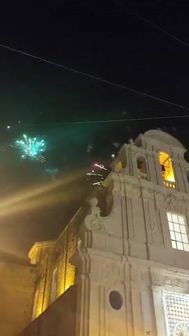 The video captures a nighttime scene with fireworks exploding in the sky over a stone building, possibly a church, which is illuminated by yellow lights. The building features architectural details like arches, a bell tower, and ornate carvings. The fireworks range in color from green and white to pink and red, creating a dazzling display against the dark sky. Wires are visible strung across the top of the frame. The camera angle is low, looking up at the building and the fireworks. The video is likely a recording of a celebration or event involving fireworks.