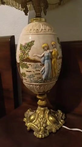 The video shows a close-up, eye-level view of a decorative lamp. The lamp has a large, ovular base with a creamy white finish. The base is adorned with a bas-relief scene featuring two women in flowing dresses, one in blue and the other in yellow, standing in a landscape with trees and water. The base is supported by an ornate, gold-colored pedestal. Above the base, the lamp transitions to a metal structure that holds a lampshade. The lampshade is a creamy, textured material, possibly fabric or faux leather, with a decorative fringe hanging from its bottom edge. The lighting in the scene is dim, suggesting an indoor setting, possibly a home. The lamp is placed on a dark wooden surface, which could be furniture like a table or dresser. The background is blurred but appears to be a wall and possibly another piece of furniture. There are no visible people actively interacting with the lamp. The camera briefly pans slightly to the right and then left, offering a more complete view of the lamp's design.