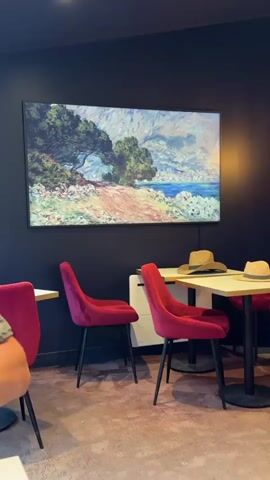 The video pans across a room with dark walls and a textured floor. Three plush red chairs are visible, two facing a table with two straw hats on it, and one in the foreground. A large framed painting hangs on the wall, depicting a scenic landscape with trees and water. The painting changes to another artwork, this one showing a woman and a child sitting amongst roses. A white radiator is situated below the paintings.