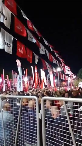 Here is a detailed description of the video:

The video shows a large crowd of people gathered at night, likely for a political rally or event. The crowd is dense, with people standing close together behind metal barriers. Overhead, strings of red and white flags with the letters "CHP" are stretched across the area. Many people are looking toward the front of the crowd, possibly at a stage or speaker. The scene is illuminated by streetlights and lights on tall poles. The atmosphere seems energetic, with a sense of anticipation or excitement.