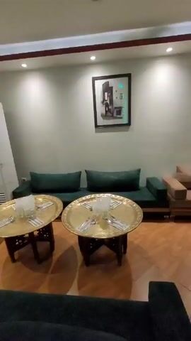 The video shows the interior of a room designed to mimic a traditional Moroccan setting. The room has low seating arrangements comprised of plush cushions and sofas in various neutral tones, and intricately carved round tables made of what appears to be brass. The floor is covered with wooden tiles arranged in a geometric pattern. Several ornate hanging lamps provide ambient lighting, and decorative sconces are mounted on the walls. Niches in the walls display small ornaments. Artwork adorns the walls, and curtains with decorative trim cover the windows. The overall impression is of a cozy, private dining or lounge area, likely within a restaurant or hotel.