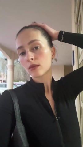 A woman with fair skin, dark hair in a bun, and wearing a black long-sleeved zip-up shirt stands beneath an archway with pillars. She has silver hoop earrings and a silver bracelet. She appears to be outdoors, possibly in a parking area, as cars are visible in the background. The sky is a dusky blue. The woman briefly holds her hand to her ear, then makes a gesture with her fingers, and she looks directly at the camera.  An Islamic call to prayer can be heard.