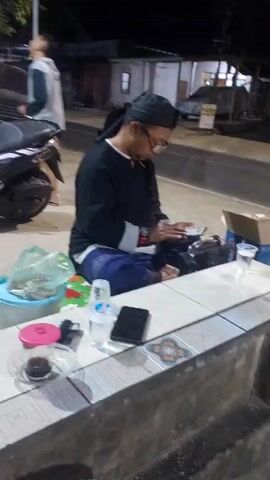 A man in a black head covering and glasses sits cross-legged on a tiled surface, looking at his smartphone. He wears a black shirt with white trim on the cuffs and a blue patterned sarong. A small cup of dark liquid sits on a saucer to his left, next to a lighter and a small pink-lidded container. To his right are two plastic cups, one with clear liquid and a bottle of water, and another smartphone. Behind him, a motorcycle is parked. The scene appears to be outdoors at night, with a road and buildings in the background. Vehicles are seen driving by in the background.