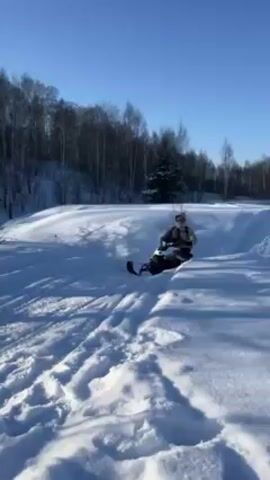 A person wearing a white jacket and carrying a gray backpack is riding a white and black snowmobile on a snow-covered path near a wooded area on a sunny day. The snowmobile makes tracks in the snow as it goes around a bend and then makes a sharp turn to the left. The person stops the snowmobile, ending the video.