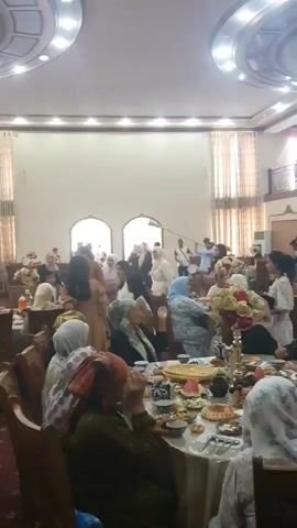 The video shows a gathering of people inside a brightly lit hall with ornate ceiling features and draped curtains. In the foreground, several people are seated at tables laden with food. The camera pans towards the back of the hall where a group of musicians are playing. One musician is blowing into a long, horn-like instrument, while another plays a drum. Several people are standing and interacting in the background, and some are dancing. The overall atmosphere appears to be festive, possibly a celebration or cultural event.