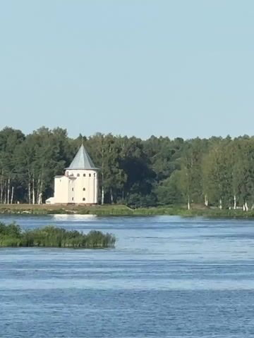 The video opens with a still image of a white, conical-roofed building with arched windows situated on the edge of a body of water. Tall green trees form a dense forest behind the building. The water in the foreground is a calm blue, with some reeds visible along the bank. The sky above is clear and a pale blue. The scene appears to be a landscape photograph.