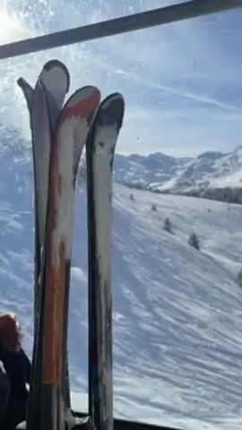 The video shows a view from inside what appears to be a ski lift or gondola. Three pairs of skis are visible, held in a rack inside the lift. The skis are different brands and colors, covered with some snow. Outside, there's a snowy mountain landscape under a blue sky with wispy clouds. The slopes are visible, with some scattered trees. A hand is visible, briefly touching the skis. Light flares from the sun are visible on the window.