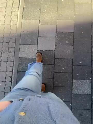 The video shows a person walking on a paved sidewalk. The person is wearing blue jeans, brown shoes, and a blue, fuzzy cardigan with gold buttons. The camera is positioned at the person's chest, looking down at their feet and the sidewalk. The sidewalk is made of gray paving stones arranged in a rectangular pattern, with a border of smaller, textured paving stones on the left. There is dappled sunlight and shadows on the sidewalk.