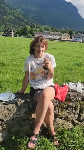 Here is a detailed description of the video:

A young woman with auburn hair in a ponytail is sitting on a rough, stone wall. She's wearing a white t-shirt with a graphic design, black shorts, and black sandals. Her legs are crossed at the knees. She has tattoos on her arms and legs. She raises a glass of a dark-colored drink, drinks from it, and then lowers the glass while gazing around. To her left on the wall is a plate, a knife, and an empty glass. To her right is a red bag and a folded white cloth.

The background features a bright green field, houses, and majestic mountains under a clear blue sky with a few wispy clouds. The scene suggests a peaceful, outdoor setting, possibly during a picnic.