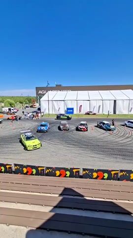 Several cars are performing drifts on a paved track. The track is circular with several orange cones placed around it. In the background, there is a large white tent with "AQUACENTER" written on the building behind it. There are spectators in the background and a few people standing near the cars. The sky is clear and blue.