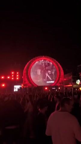 A large crowd of people are gathered at an outdoor event at night. The scene is dominated by red lighting and a large, circular screen in the background. On the screen, two individuals appear to be performing or DJing. The crowd is densely packed, with many people facing the screen. Some individuals are holding drinks, and some are wearing hats. The overall atmosphere appears to be a concert or festival.