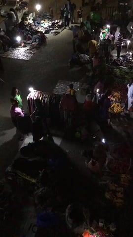 The video shows a busy outdoor market at night. People are gathered around stalls displaying various goods, including clothing and produce. Several individuals are using flashlights or their phone lights to illuminate the merchandise. Some people appear to be browsing, while others are engaged in transactions. A few vehicles are visible in the background, their headlights cutting through the darkness. The overall atmosphere suggests a vibrant and active marketplace.