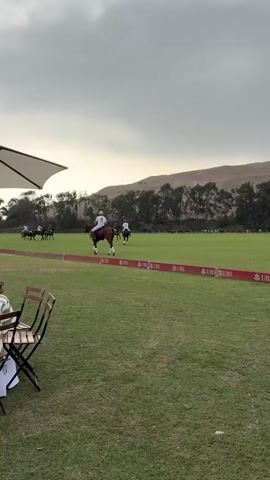 The video shows a polo match on a green field. Several horses and riders are visible on the field, some of whom are actively playing polo. There are also spectators at tables and chairs in the foreground, along with an umbrella. In the background, there are trees and hills. The sky is overcast. The red ribbon along the field has the text "UBS" printed on it repeatedly.