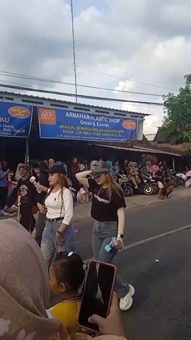 The video shows a parade taking place on a street in front of a store called "ARMAHAM PLASTIC SHOP." The store is advertising various plastic products like cups, boxes, and other food-related supplies. A crowd of people is gathered on the sidewalk to watch the parade.  Several women in traditional clothing are marching down the street. They are wearing black tops and patterned skirts. The sky is blue with some clouds. Someone is recording the parade on a smartphone, with a small child standing in front of them.