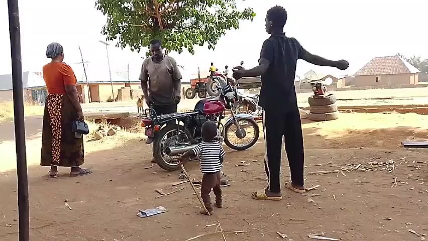 A medium shot captures a dusty yard in a rural setting. In the foreground, a woman in an orange top and patterned skirt stands to the left, holding a purse. A young child in a striped shirt stands in the center, facing a red motorcycle that a man is working on. Another man in black pants and a black shirt stretches his arms above his head to the right of the motorcycle. 

Behind them, a green tree provides partial shade. In the background, there are buildings, a tractor, and livestock visible in the distance. The ground is covered in dirt, with metal debris scattered around the motorcycle.