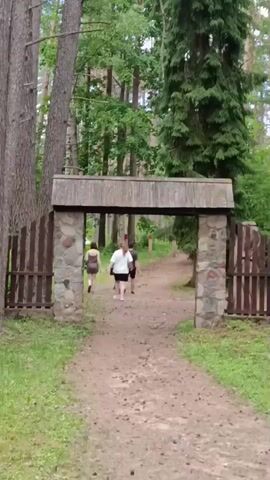 This is a video of a dirt path leading through a forest. There is a wooden gate with stone pillars. Three people, two women and a man, are walking away from the camera through the gate. The path is surrounded by trees and grass.