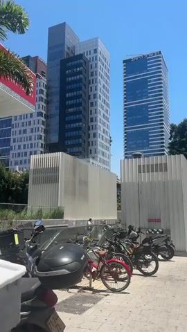 The video pans across an urban scene under a bright, clear sky. In the foreground, several bicycles are parked, one of them red. To the left, the corner of a black scooter's storage compartment is visible. Behind the bicycles, there are modern, light-colored structures with a corrugated facade. In the background, tall office buildings with glass and metal exteriors dominate the skyline. One building is particularly striking with its blue-tinted glass and white horizontal accents. A few trees are interspersed between the buildings. To the right, a paved sidewalk runs alongside a street with moderate traffic. Cars, including a silver sedan and a white SUV, are driving on the road. Two people are walking away from the camera on the sidewalk. Two Israeli flags are affixed to a lamppost.