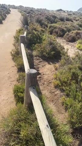 A dirt path winds uphill through a landscape of sparse vegetation and sandy terrain. A rustic wooden fence lines the right side of the path, made of simple posts and horizontal rails. The vegetation appears to be dry grasses and low-lying shrubs, typical of a coastal or arid environment. In the distance, a small red flag is visible at the top of the hill. The sky is bright and clear, suggesting daylight. The camera appears to be moving along the path, giving the viewer a first-person perspective of the ascent.