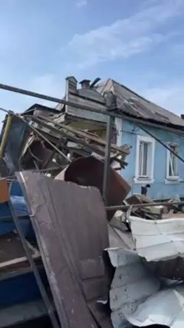 Here is a concise description of the scene in the video:

The video shows the aftermath of significant destruction. The scene centers on a severely damaged house, with its roof and structural elements largely collapsed and intermixed with other debris. Items like metal sheeting, furniture (cabinets or boxes), and various construction materials are visible amidst the wreckage. The house appears to be light blue with white-framed windows. The sky is partially clear, and a few trees can be seen in the background. Overall, the video depicts a scene of substantial damage, likely due to an explosion, severe weather, or a similar destructive event.