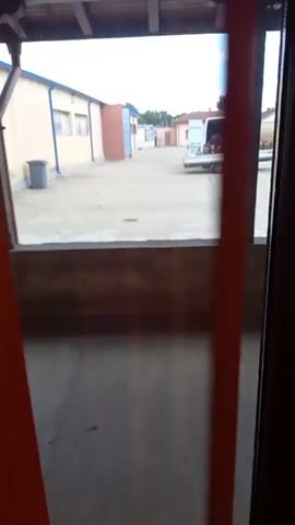 The video shows an outdoor courtyard with several buildings. In the foreground, there is a wooden structure that appears to be a porch or covered area. Through the opening of this structure, the courtyard is visible. The courtyard is paved and appears to be somewhat dusty. There are multiple buildings surrounding the courtyard, some of which are industrial in appearance with light-colored walls and windows. A small grey trash can is visible near one of the buildings. Further into the courtyard, there is a white truck with its back doors open, and a person is visible inside or near the back of the truck. Another white van is parked in the distance. The overall impression is that of an industrial or commercial outdoor space, possibly a loading dock or storage area. The lighting suggests it is daytime, but the sky is overcast.