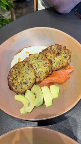 The video shows a close-up, overhead shot of a plate of food. The plate is a light brown color with a slightly textured rim. In the center of the plate are four round, golden-brown fritters, which appear to be made with vegetables like broccoli and quinoa, as visible green specks and small seeds are mixed throughout. To the left of the fritters is a dollop of white sauce, possibly yogurt or sour cream, with a drizzle of orange oil on top. To the right of the fritters are three slices of smoked salmon, curled slightly. In the lower left of the plate, there are several slices of bright green avocado arranged in a fan shape. The food is set on a dark gray surface, possibly a table, with a hint of a green plant in the upper left corner and a person's legs and feet in the upper right, suggesting an indoor dining setting. The lighting is soft and natural, highlighting the textures and colors of the food.