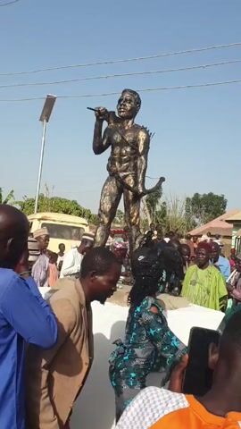 A statue of a man is in the center of a crowd of people. The statue is of a man with a spear in one hand and a bow in the other. He is wearing a loincloth and has a smile on his face. There are two dogs at his feet. The statue is on a white pedestal. People in the crowd are taking pictures of the statue with their phones. A light post is visible to the left. There is a yellow van and some buildings in the background. The sky is blue.