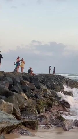 A group of people stand on a rocky pier overlooking the ocean. The sky is cloudy and a wave crashes against the rocks. The people are spaced out, some standing and some sitting. One person is waving.