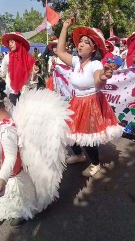 The video captures a daytime outdoor procession or parade. In the foreground, a woman in a white top, a red and white ruffled skirt, and a red hat is dancing and raising her right arm. To her left, a child is visible wearing a white costume with large white feather-like wings and a red and white outfit. Further back, other individuals, including women in red hijabs and hats, are participating in the parade. A banner with Indonesian text and graphics is visible in the background, along with trees, umbrellas, and what appears to be a motorcycle. The atmosphere suggests a festive or celebratory event.