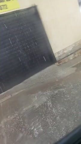 The video shows a black metal gate in the background with a yellow sign on the upper left corner. The gate is partially obscured by rain. In the foreground, the ground is wet and reflecting light, indicating it is raining. Small droplets of water are visible falling. The overall scene appears to be outdoors during a rain shower.